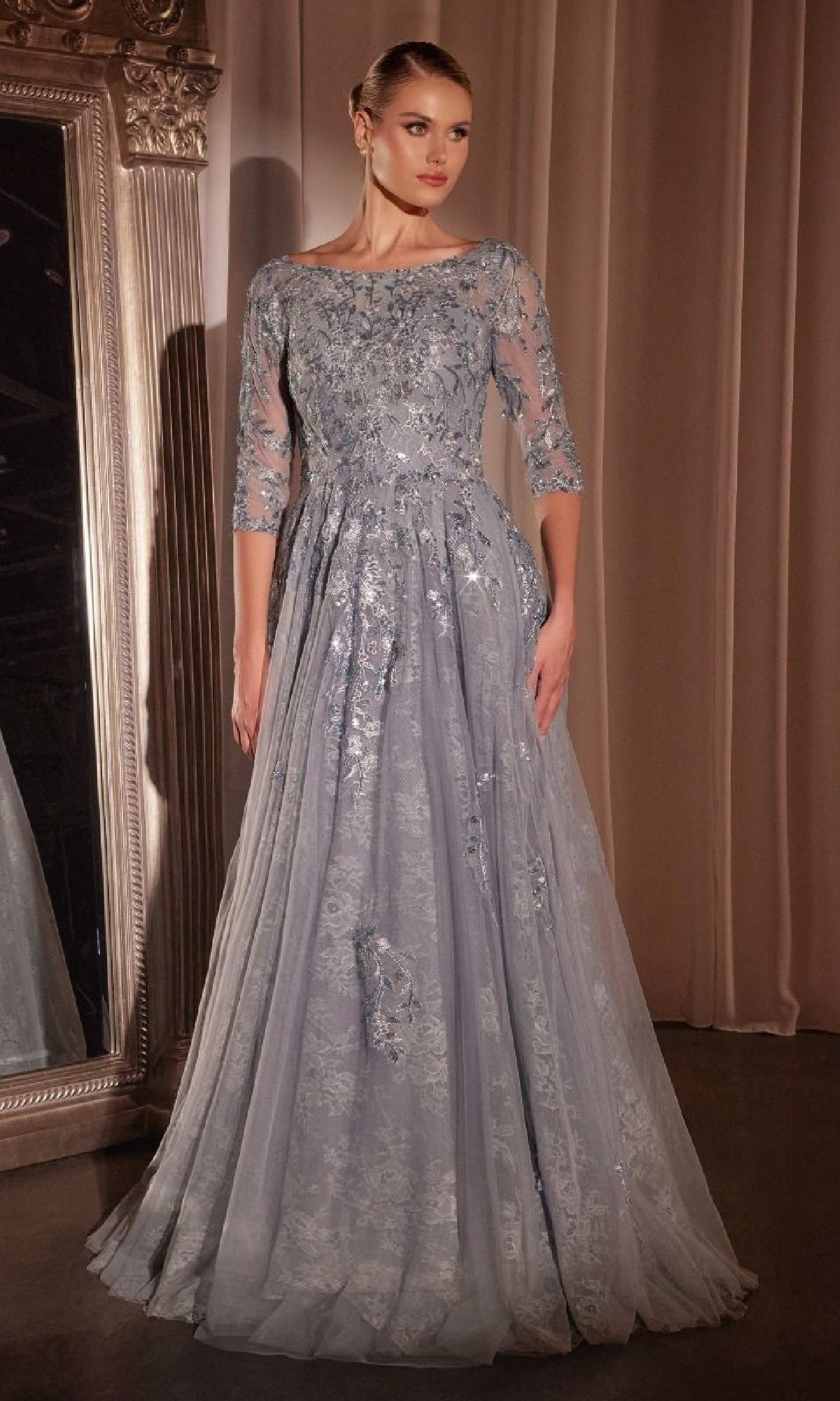 A woman wearing the Long Sleeve Blue Lace Formal Gown—Ladivine CK2401 stands beside a large ornate mirror and beige curtains.